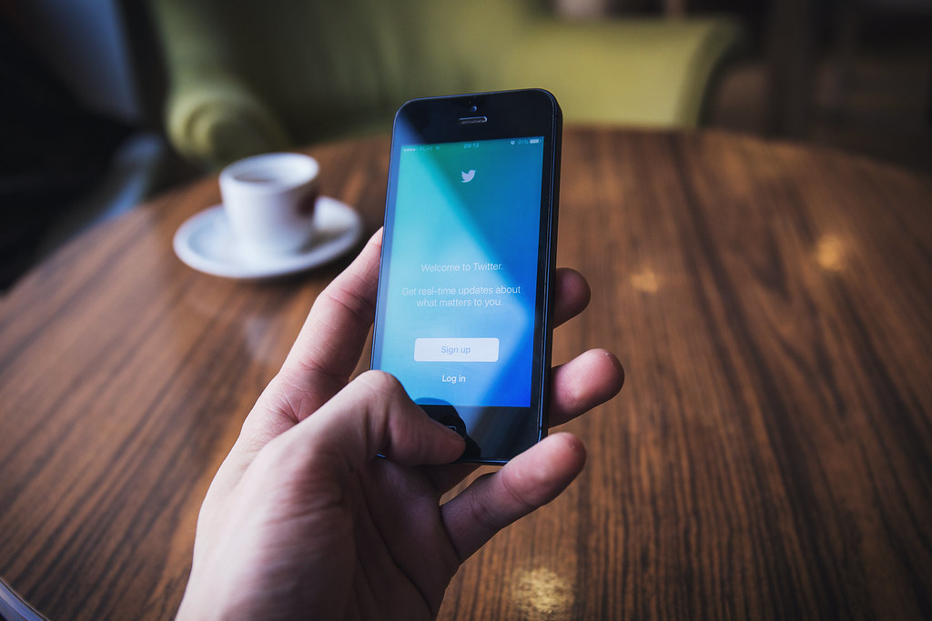 A hand holding a smartphone showing a social media app while sitting at a table